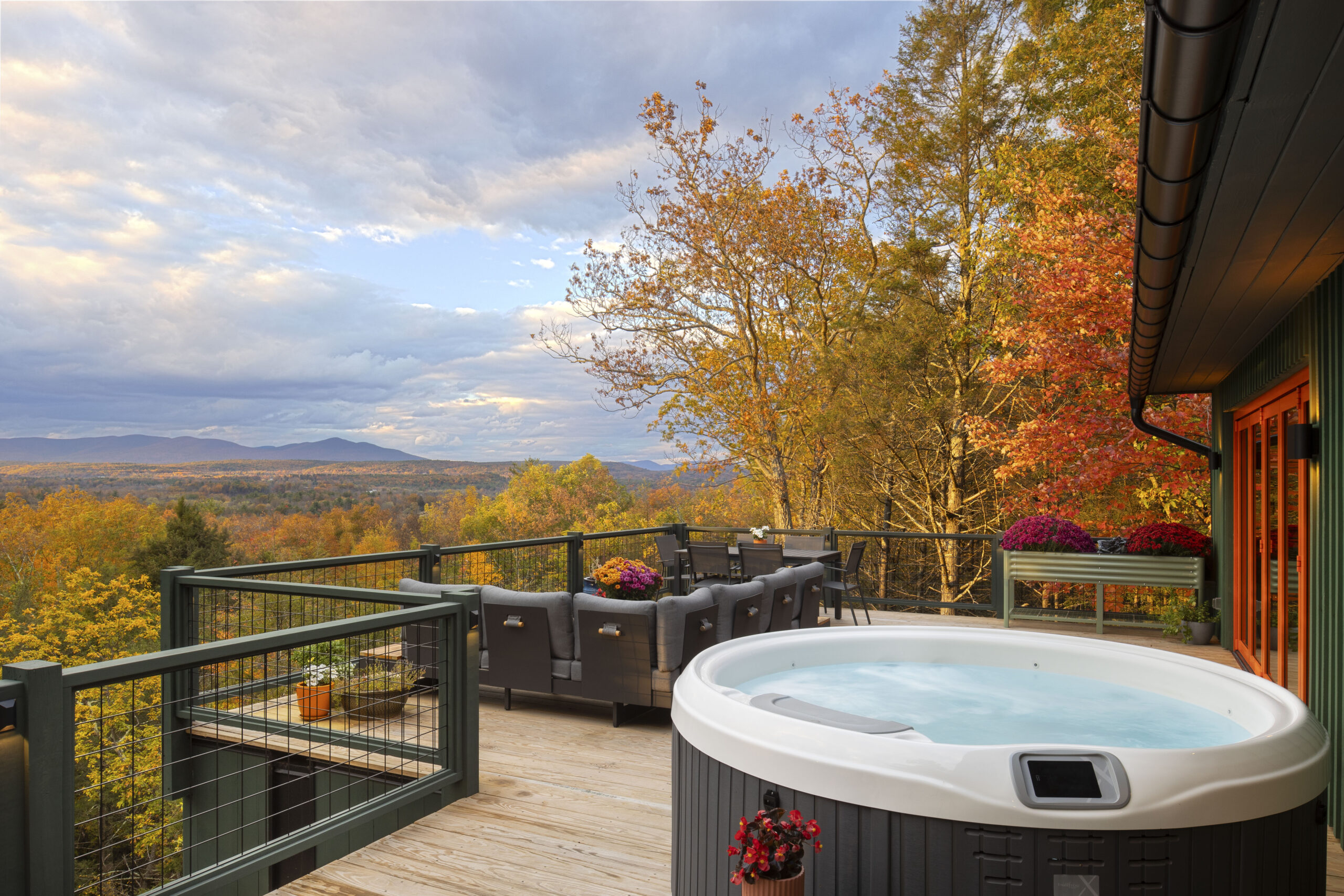 A wide-angle shot of the wooden upper deck featuring a large, round grey hot tub. A dark grey sectional sofa and a dining area are positioned to take in the sprawling view of the autumn forest and distant blue mountains. Red floral planters on the deck mirror the vibrant fall foliage in the background.