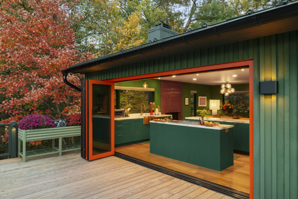 A wide view of the kitchen from the wooden deck through large, red-framed bifold doors that are fully open. The kitchen features forest green cabinetry, white marble countertops, and a large central island. The green of the cabinets perfectly echoes the lush forest trees just outside.