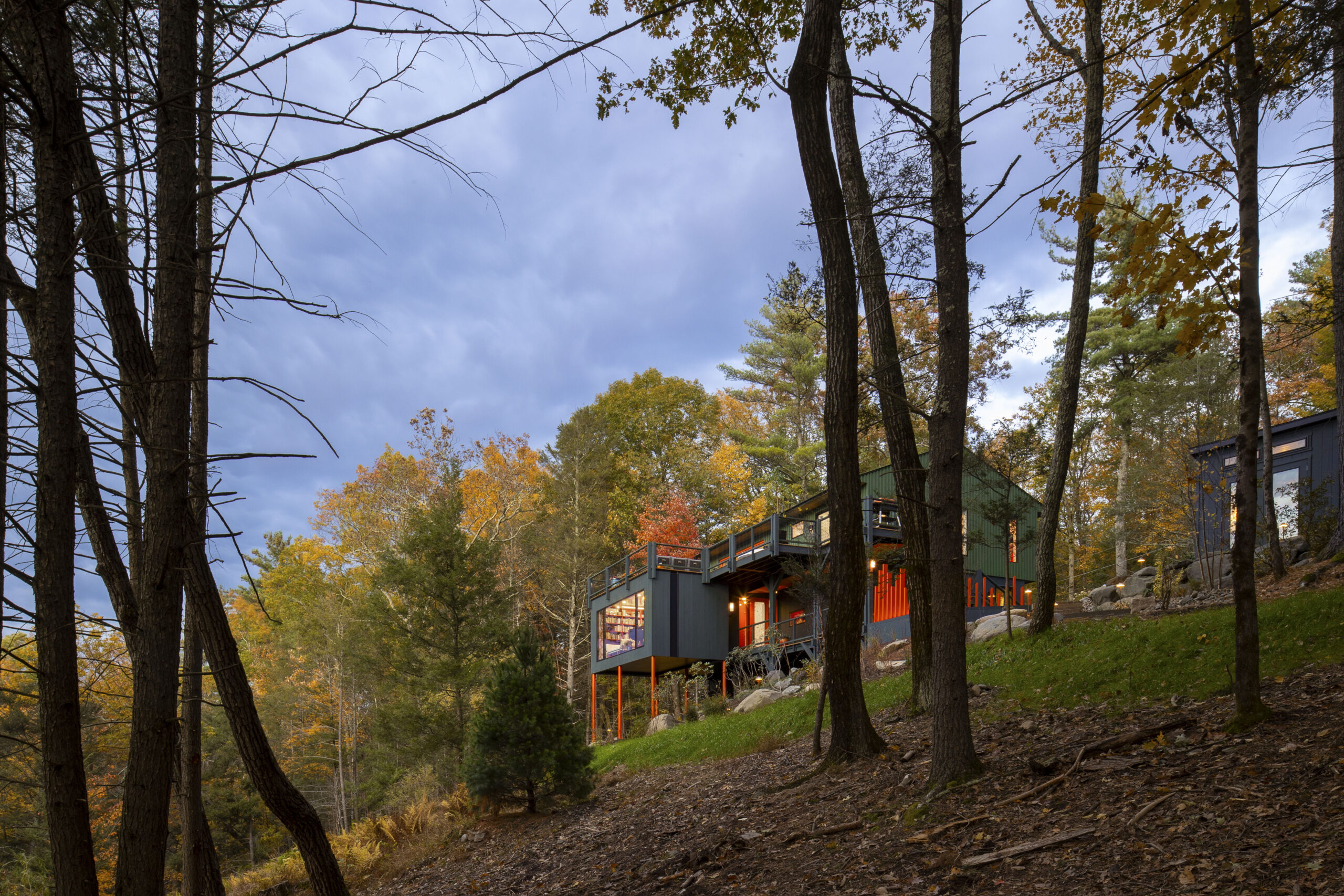 A low-angle view taken from the bottom of a wooded slope looking up at the house. The dark trunks of foreground trees frame the modern structure, which sits perched on the hillside. The orange pillars and fence pop against the green grass and the moody, overcast sky.