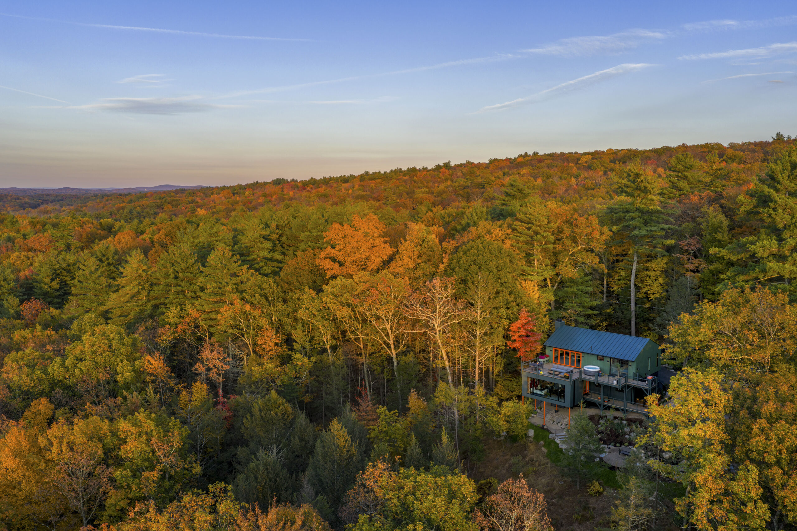 A distant aerial view showing the house nestled into a vast, rolling forest of autumn colors under a clear blue sky. The house appears as a small, modern jewel within the dense Hudson Valley canopy, highlighting its seclusion and the way the dark green siding camouflages the structure among the pines.
