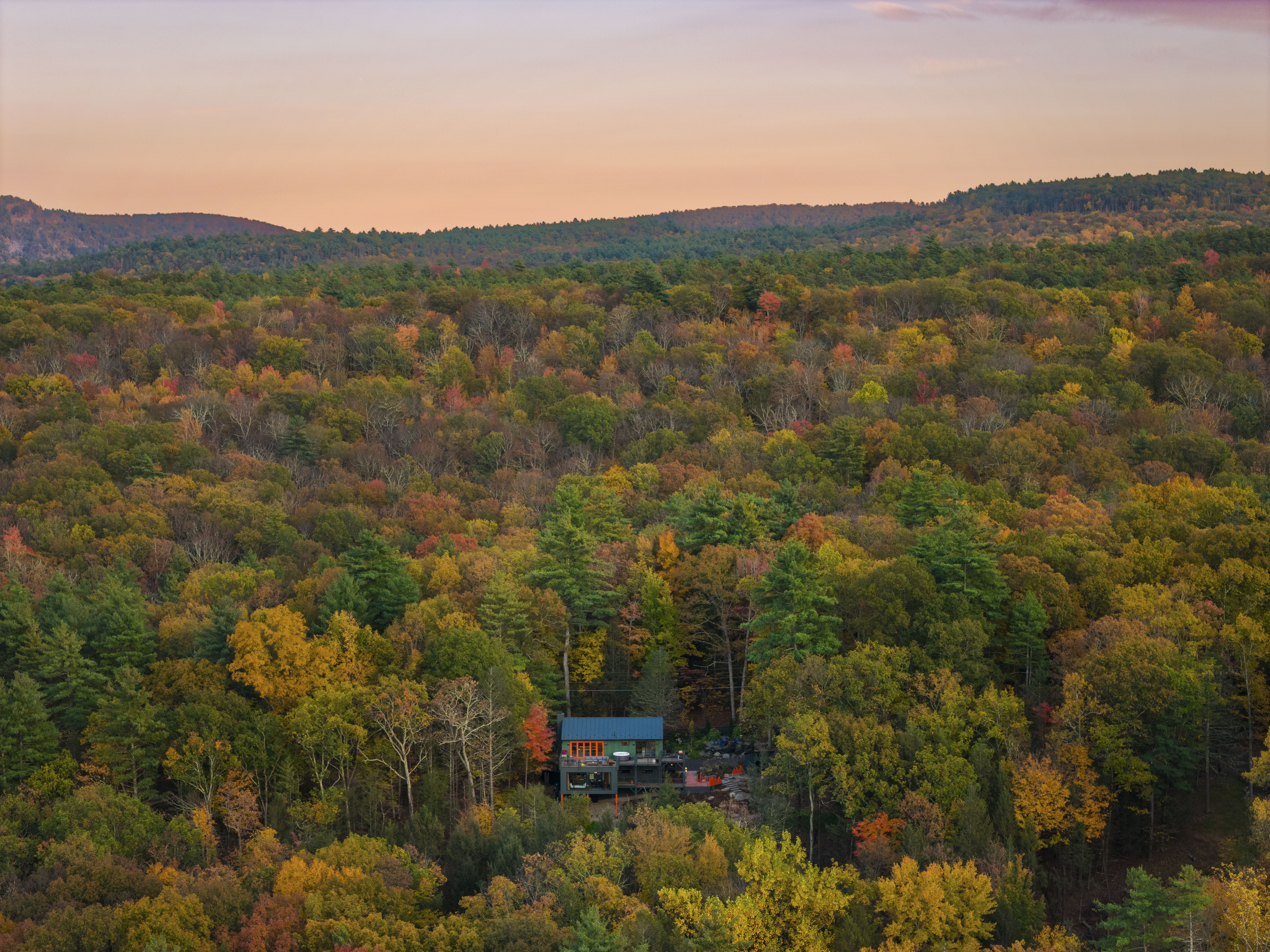 A wide landscape shot at dusk with the sky fading from soft blue to a warm peach. Woodland Pop House is centered in the frame, surrounded by a sea of colorful treetops. The distant mountains of the Hudson Valley create a layered silhouette on the horizon.