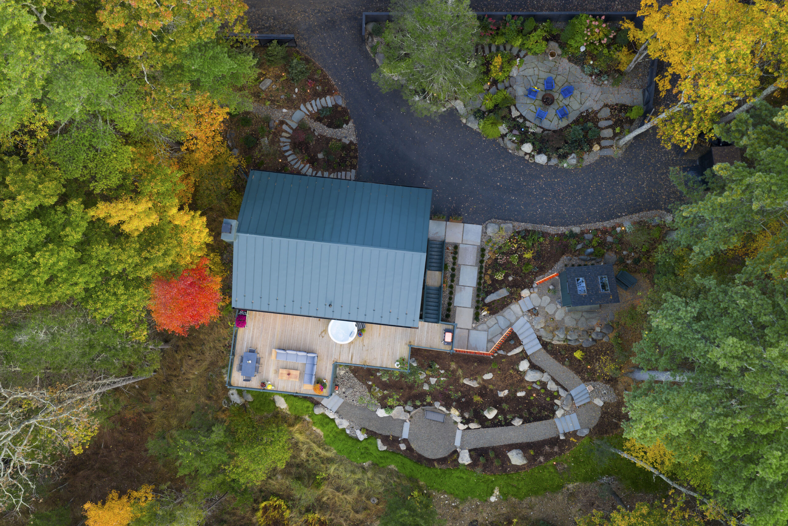 A top-down aerial photograph of the Woodland Pop House and its grounds during autumn. The dark green metal roof stands out against a tapestry of orange, yellow, and red trees. The image reveals the home’s layout, including a large wooden deck with a hot tub and outdoor lounge, a separate stone fire pit area with blue chairs, and winding gravel paths connecting the different outdoor zones.