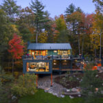 An evening view of the house’s multi-level facade. The interior lights are on, revealing a music studio with a grand piano on the lower level and a modern kitchen and bedroom on the upper level. Bright orange support pillars and an orange sculptural fence provide vivid accents against the darkening forest.