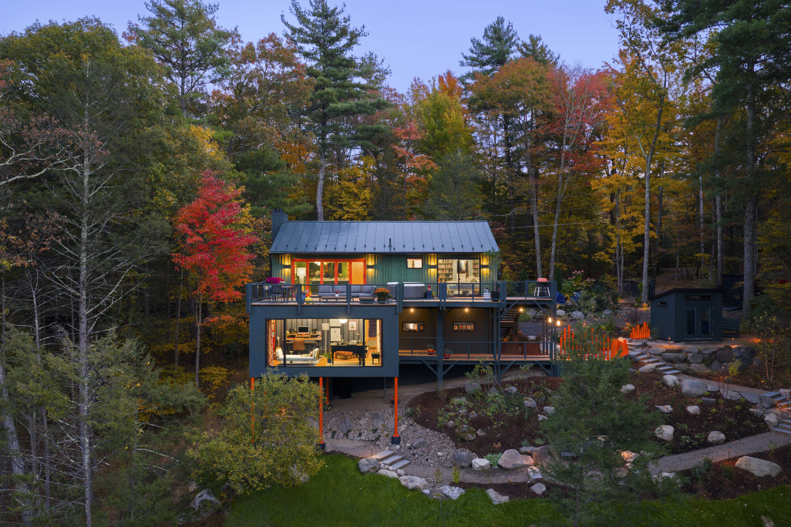 An evening view of the house’s multi-level facade. The interior lights are on, revealing a music studio with a grand piano on the lower level and a modern kitchen and bedroom on the upper level. Bright orange support pillars and an orange sculptural fence provide vivid accents against the darkening forest.
