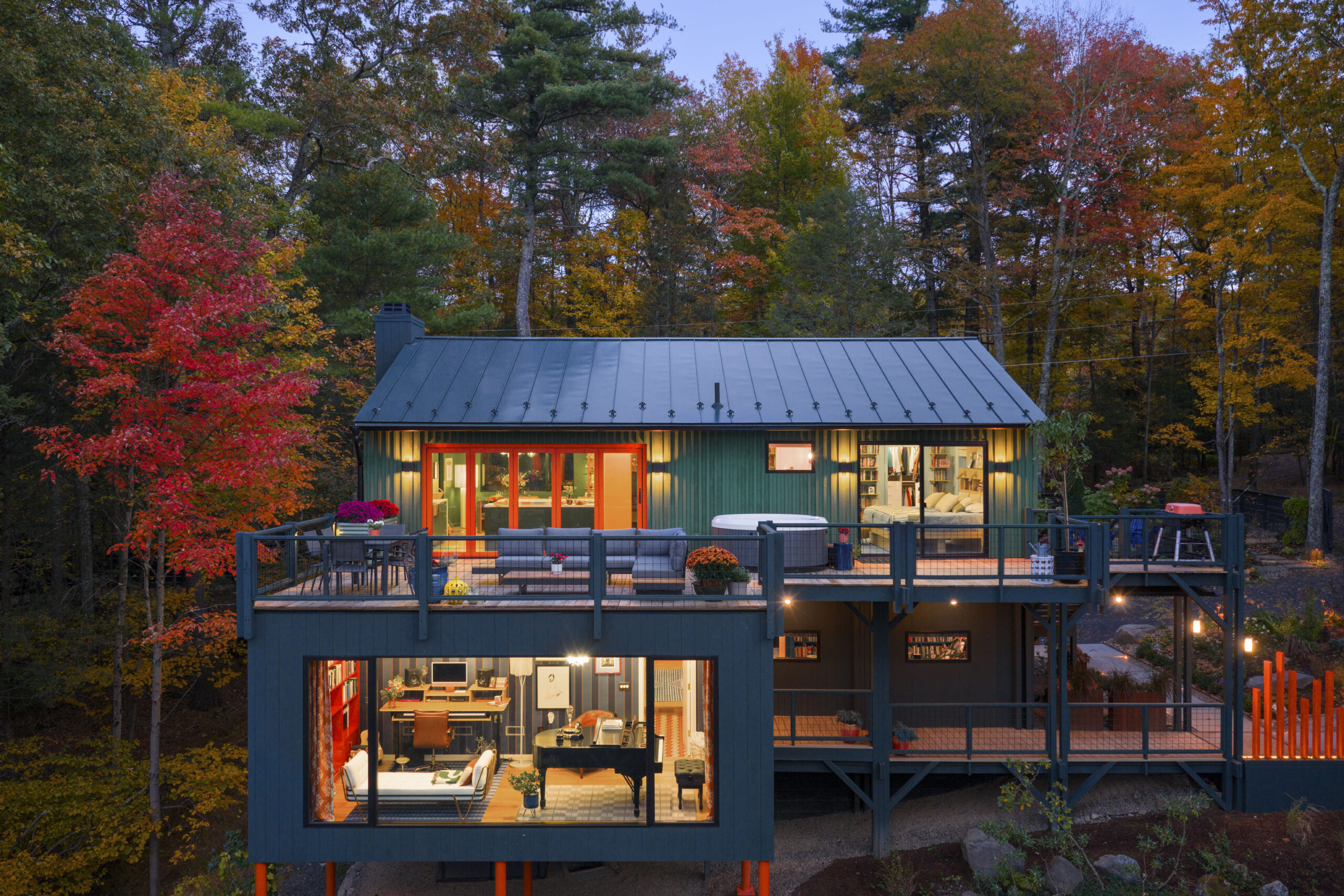 A detailed twilight shot focusing on the home’s rear elevation. The warm glow from the windows and the orange-trimmed glass doors contrast with the cool green exterior. The lower-level studio is clearly visible through large glass panes, showing a creative space filled with books, a desk, and a piano.
