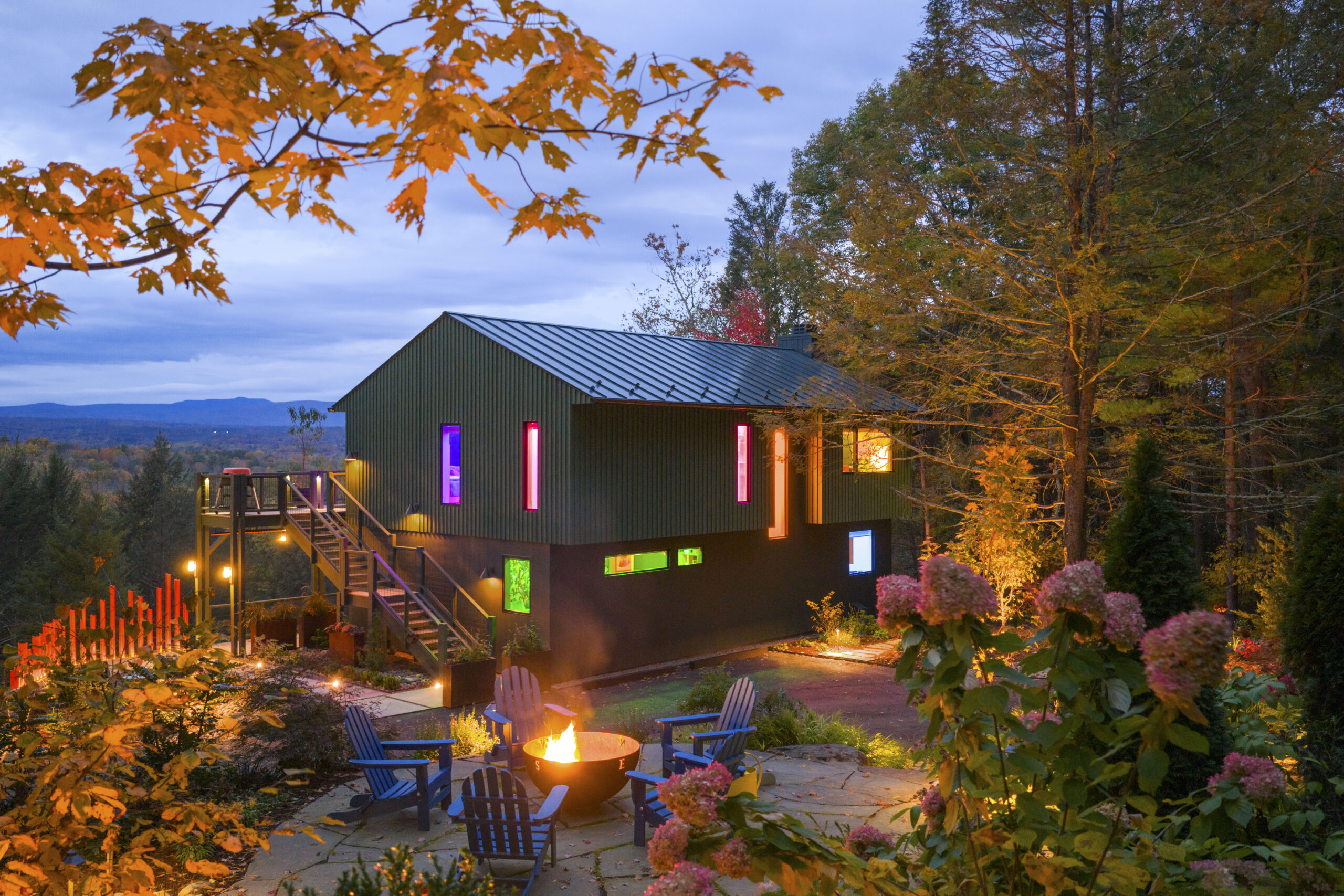 A two-story modern house at dusk, featuring dark green siding on the top and a green-grey base. The narrow vertical windows glow with vibrant neon colors—purple, pink, and orange—creating a "pop" effect. In the foreground, a stone patio features blue Adirondack chairs encircling a lit fire pit, framed by autumn maple leaves and pink hydrangeas.