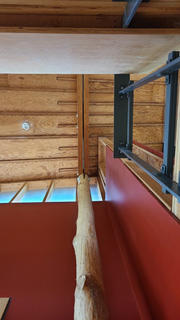 A dramatic low-angle shot showing a vertical raw wood log column meeting a modern plywood ceiling and dark grey metal framing.