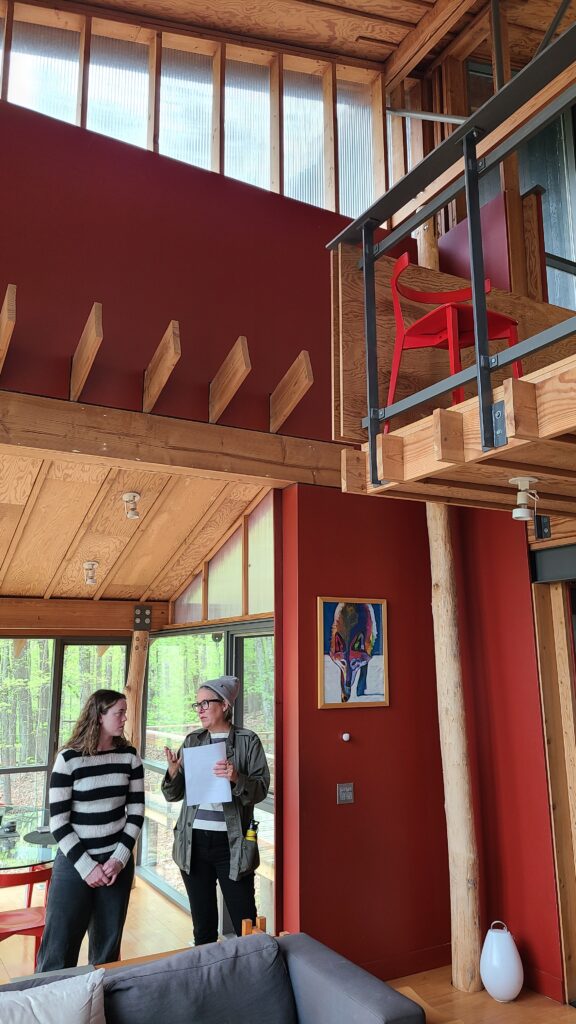 Two women converse in a modern, multi-level home featuring red walls, a natural log support column, and a bright red designer chair on a mezzanine.