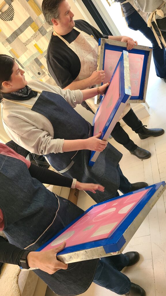 A group of people wearing aprons stand in a circle, each holding a screen-printing frame with different geometric patterns.
