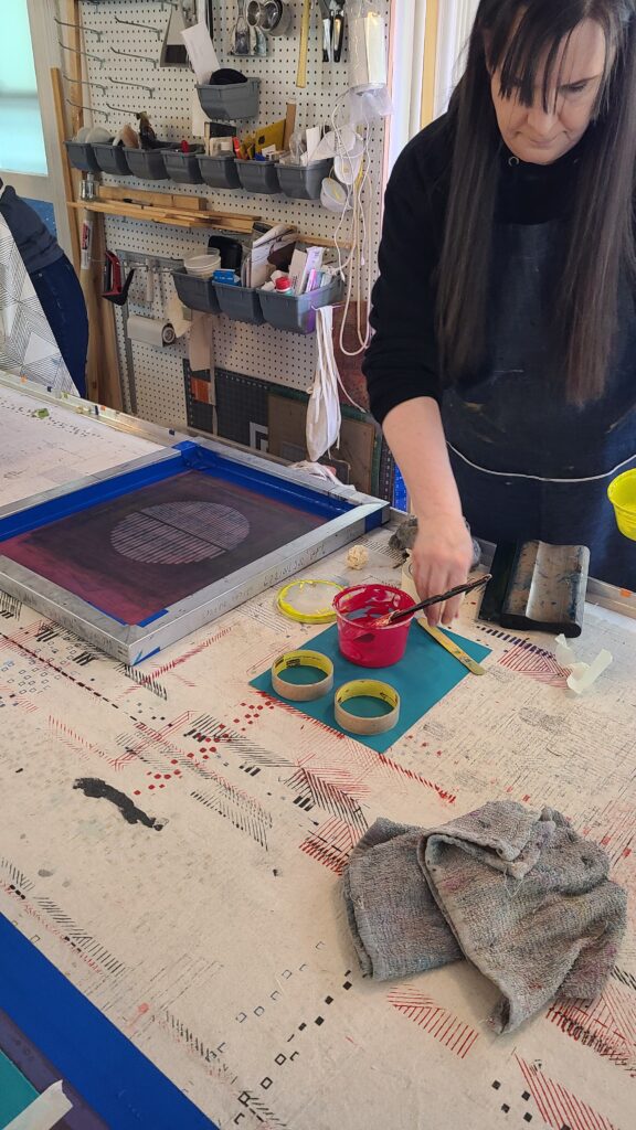 A woman in an apron prepares pink ink at a printing table next to a geometric screen-printing frame in a studio.