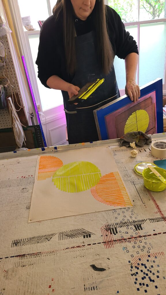 A team member holds a squeegee over a fresh fabric print of three striped, neon yellow and orange semi-circles.
