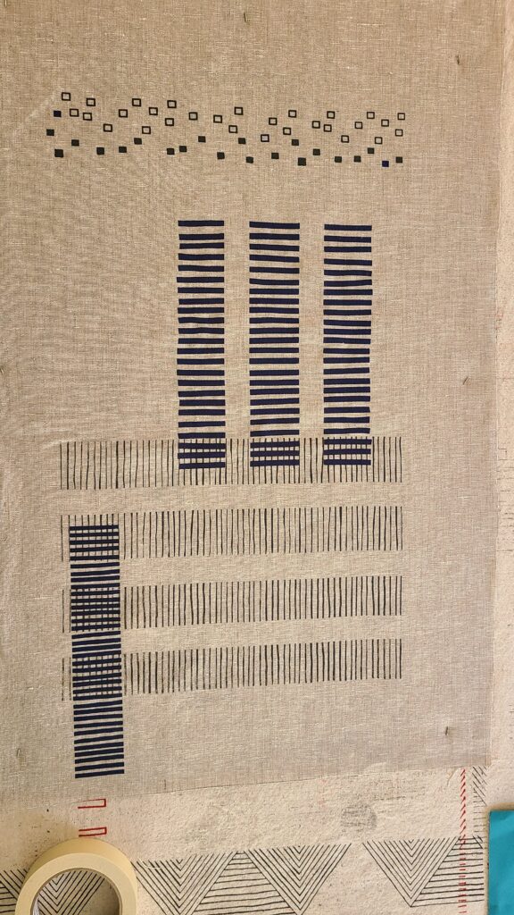 A linen fabric sample pinned to a table, featuring a dark blue geometric design of vertical bars and small square outlines.