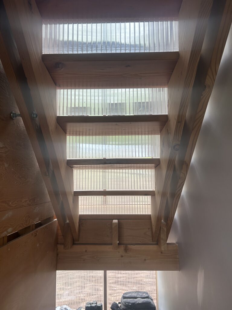 An upward view through open-tread wooden stairs, featuring translucent corrugated risers that let soft light filter through.