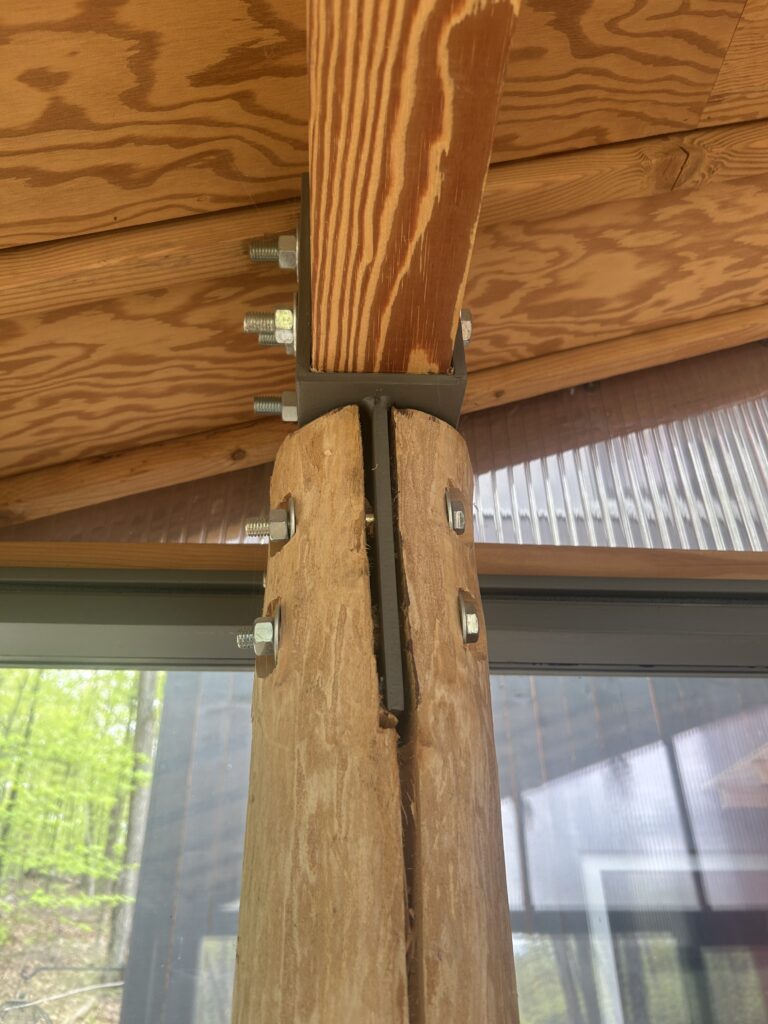 A close-up detail of a structural connection where a raw log post is bolted into a steel bracket and a finished wood beam.