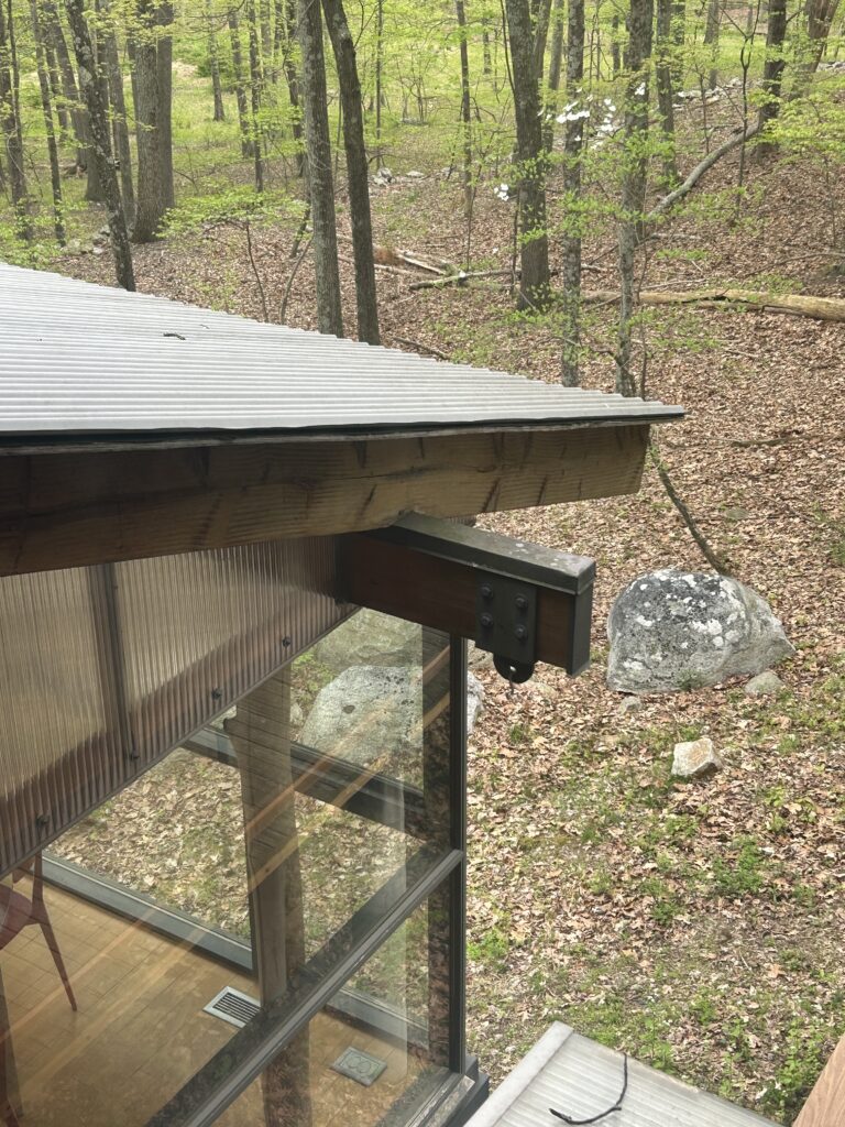 An exterior detail of a modern roof corner with corrugated metal, a dark wood beam, and a glass wall overlooking a spring forest.