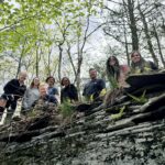 A low-angle shot of a group of seven people smiling as they peer over the edge of a mossy, layered stone wall in a sunlit forest.