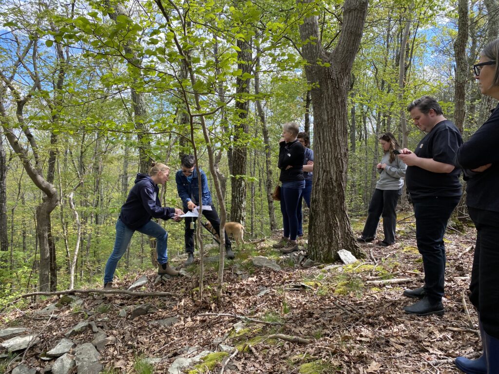 A group of people stands on a rocky, leaf-covered wooded slope, reviewing a set of site plans together under a canopy of trees.