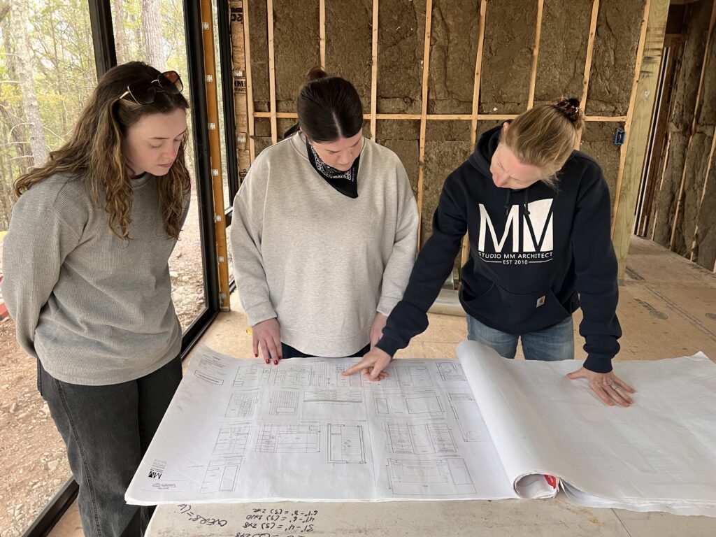 Three women review architectural blueprints inside a building under construction with exposed wood framing and large windows.