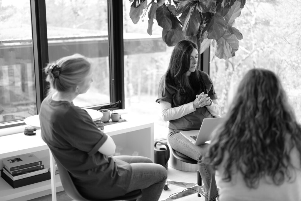 A black-and-white candid photo of three people in a meeting near a large window and a fiddle-leaf fig plant.