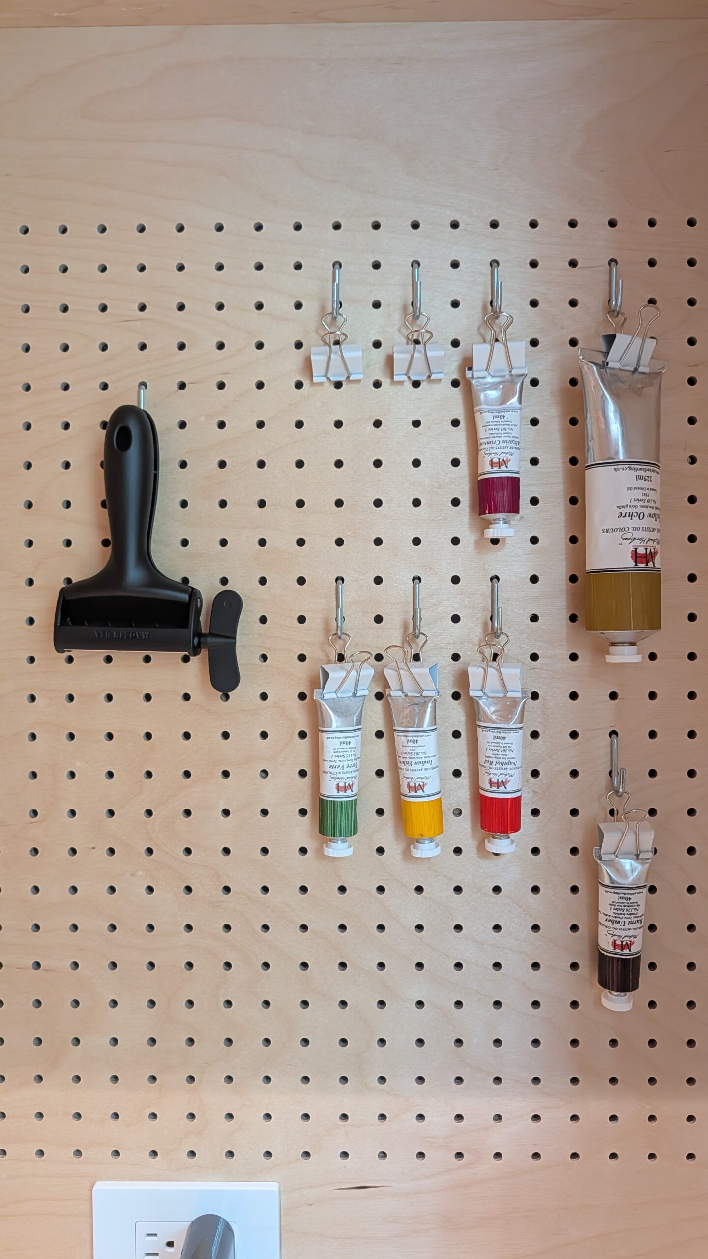 A wooden pegboard organizing art supplies, including a black brayer and several oil paint tubes secured with binder clips.
