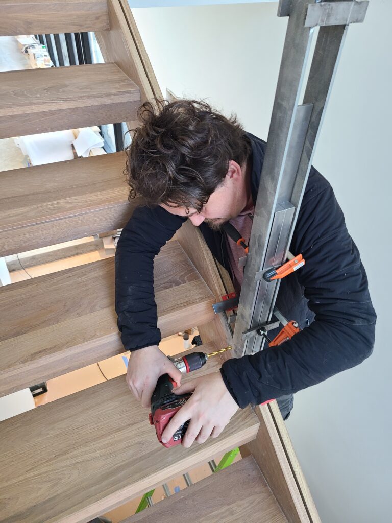 Craftsman drilling into a white oak stair tread to anchor a black steel baluster bracket