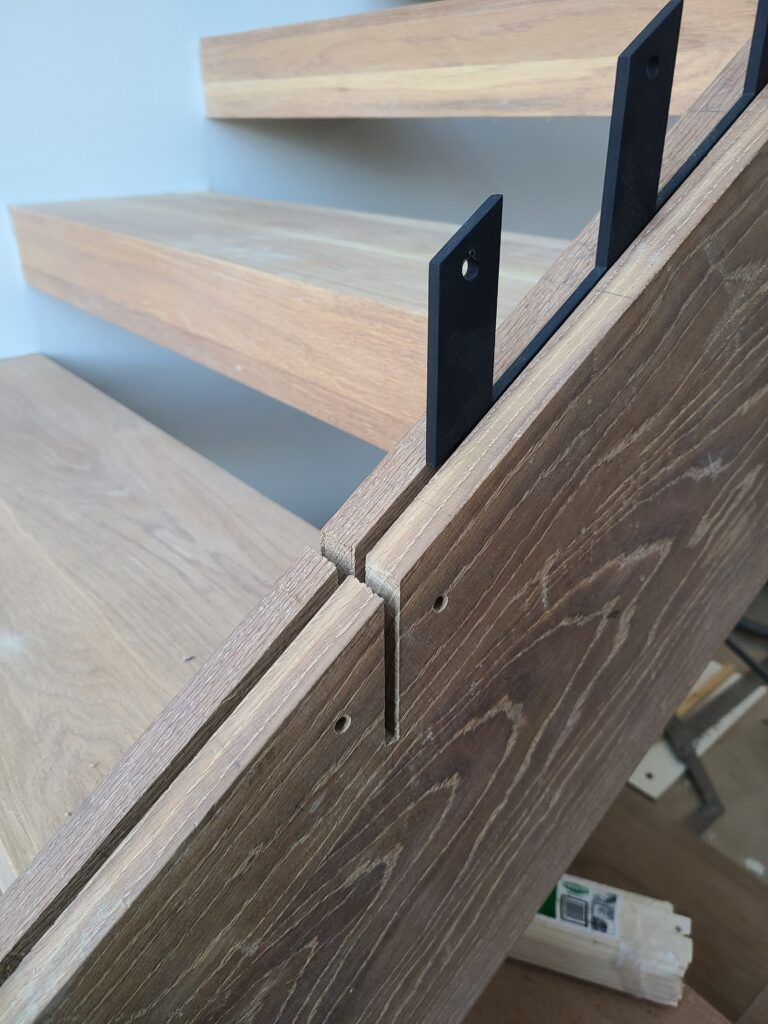 Detail of a black steel flat-bar baluster notched into a white oak stair tread