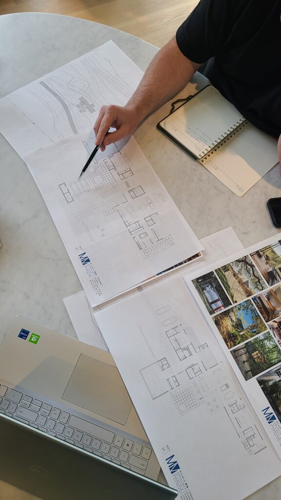 Overhead view of a hand holding a pencil over printed Studio MM Architect floor plans and a topographic site plan on a table