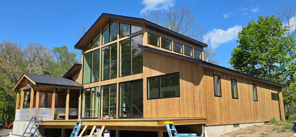 Exterior of a nearly finished modern home with tall vertical wood siding, black-framed floor-to-ceiling windows in a two-story gabled section, a covered porch area on the left, and ladders and buckets still present near the foundation.