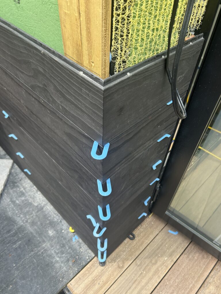 Close-up of black wood-look exterior siding corner detail held by blue plastic installation clips, next to a protected wood post