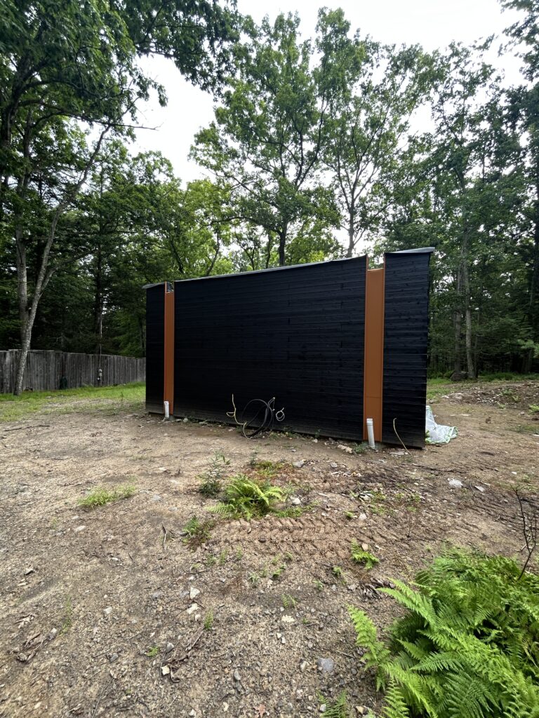 Exterior of a small black wood-clad structure with orange vertical trim accents in a wooded yard
