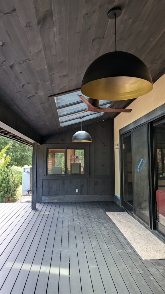 Covered outdoor porch with dark stained wood ceiling and decking, a large black dome pendant light with gold interior, and a ceiling fan