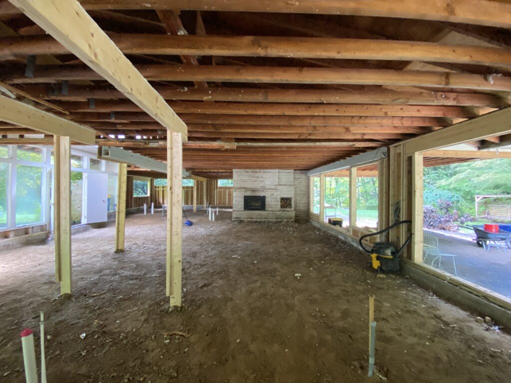 Interior of a home under major renovation with exposed floor joists overhead, dirt subfloor, wood support posts, and a stone fireplace