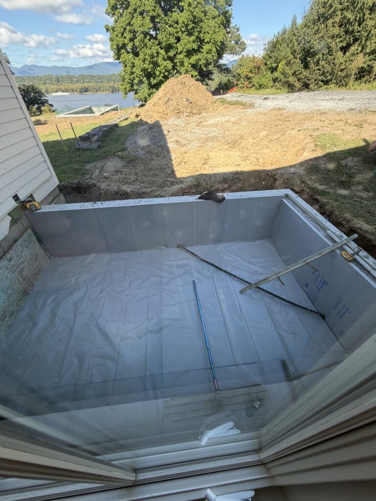 Excavated foundation lined with insulationand plastic sheeting, viewed through a window overlooking a river and distant mountains
