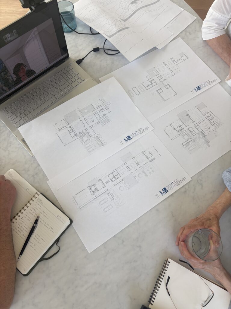 Overhead view of a table covered with four printed Studio MM Architect floor plan schemes labeled A through D, with a laptop video call open