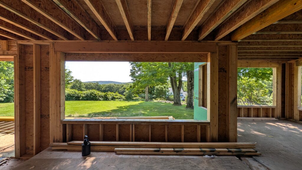 Interior view through a newly framed wall opening showing exposed wood studs, green ZIP System sheathing, and a grassy backyard beyond