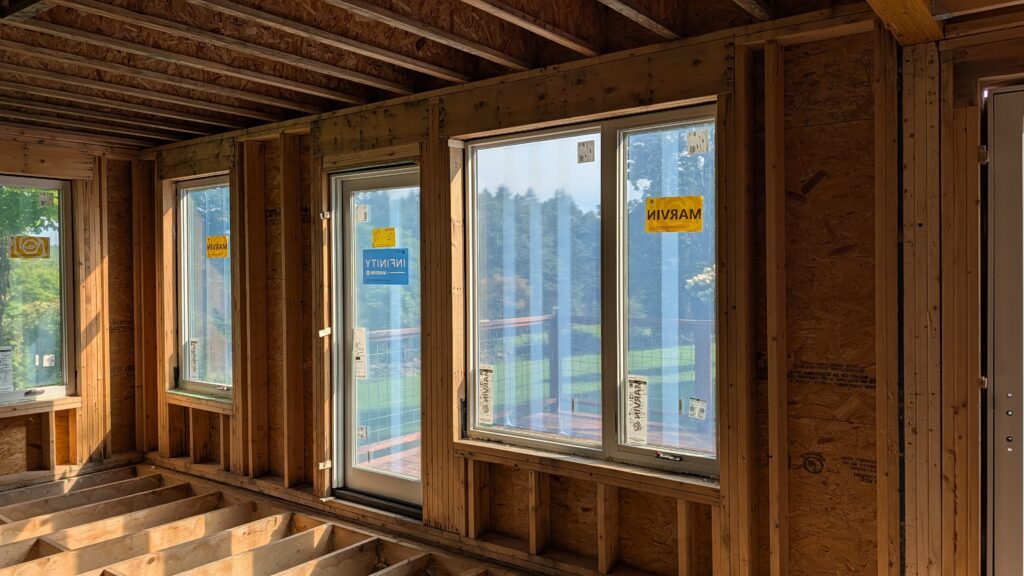 Interior of a framed addition showing exposed roof rafters and several new windows and a glass door with manufacturer stickers, overlooking a deck
