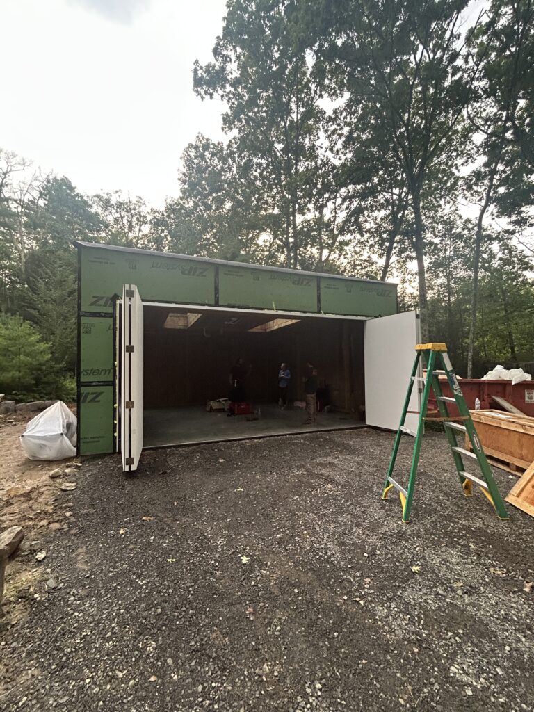 Exterior of a partially built garage or studio with green ZIP System sheathing, open bay doors, workers inside, and a dumpster nearby