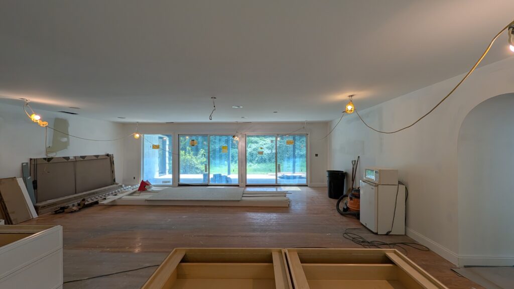 Gutted living and dining room with hardwood floors, string work-lights hanging from the ceiling, and sliding doors covered in protective film