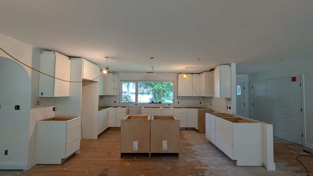 Kitchen mid-installation with white cabinetry mounted on the walls, unfinished plywood island boxes, and patchy hardwood flooring