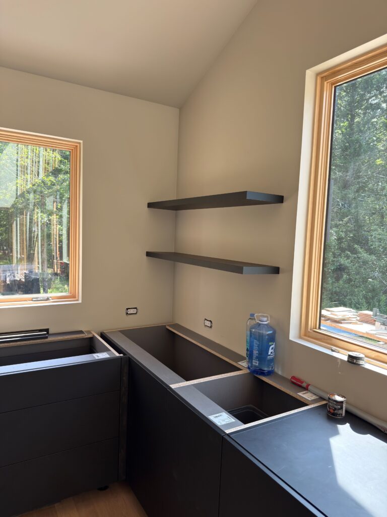 Kitchen under construction with vaulted ceiling, wood-trimmed windows, dark lower cabinetry, and a deep matte-black sink basin