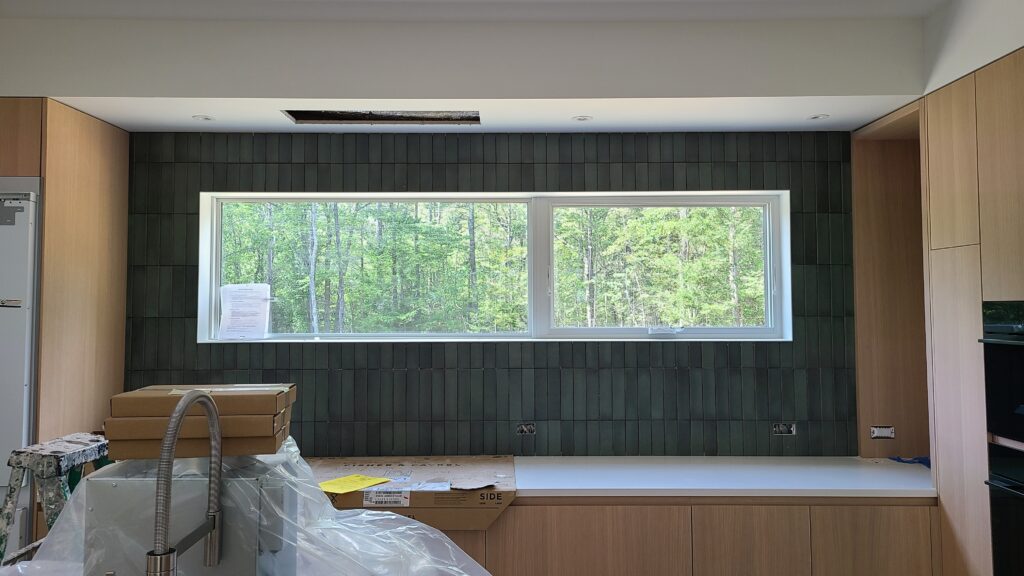 Kitchen under construction with dark green vertical-stack tile backsplash surrounding a wide window looking into forest