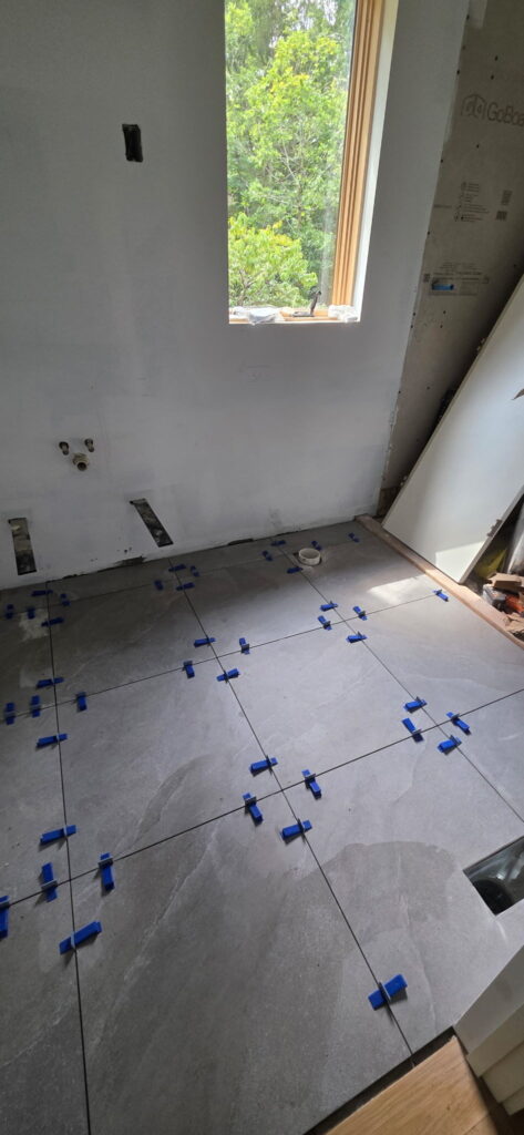 Close-up of newly laid gray tile flooring with leveling spacer clips, next to a wood-trimmed window overlooking greenery