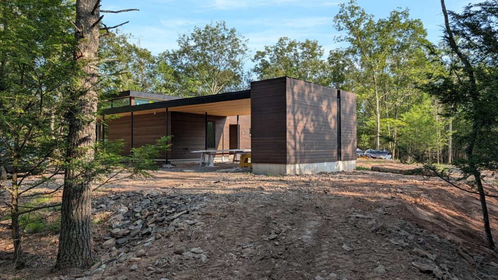 Exterior of a modern dark wood-clad house with a flat black roof and glass corner, set among trees on a rocky, partially cleared site