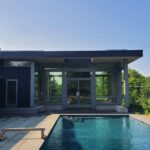 Exterior of a modern black house from the pool side, showing large glass walls, a covered patio, and a rectangular pool
