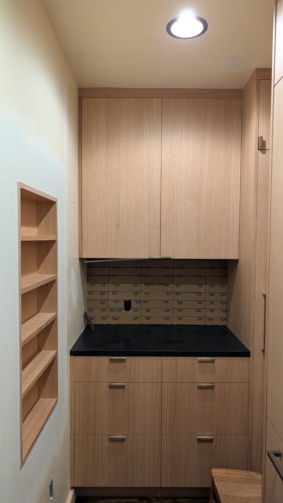 Built-in light wood cabinetry in a mudroom or pantry nook with upper and lower cabinets, black countertop, and open wood shelving