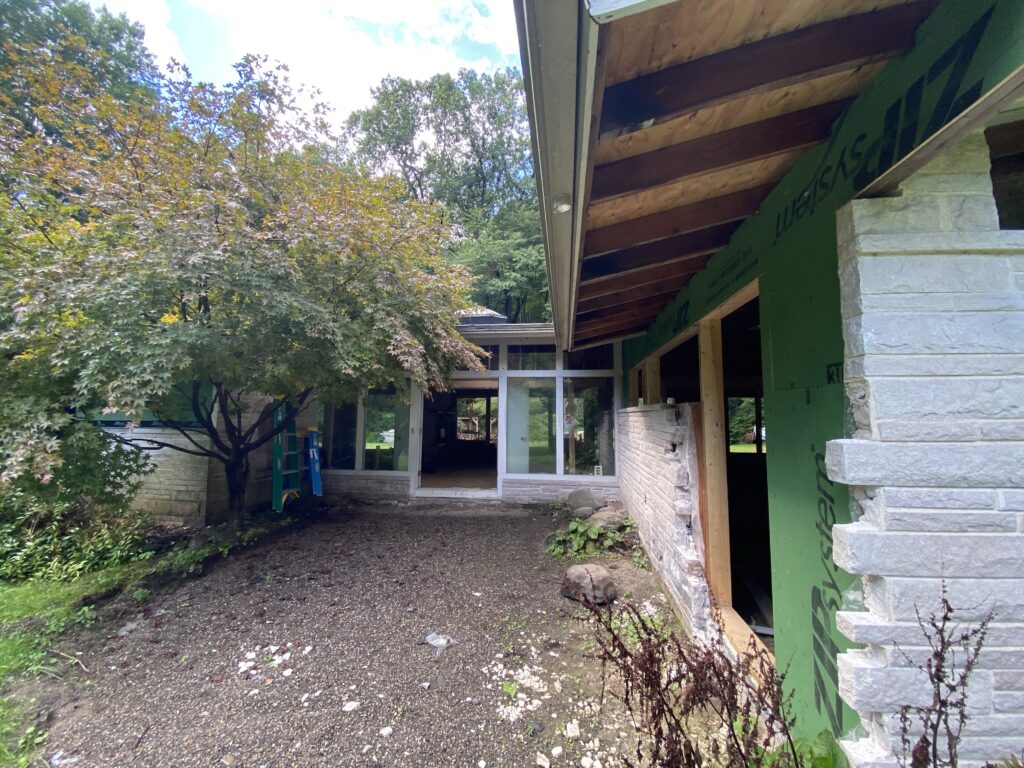 Exterior corner of a house mid-renovation showing an overhanging eave, green ZIP System sheathing, and partial stone veneer application