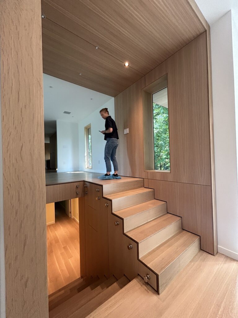 Person wearing shoe covers standing at the top of a light wood open staircase with built-in wood-paneled walls, checking their phone