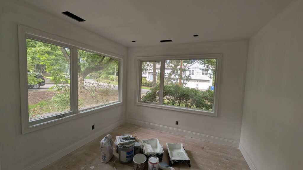 Freshly painted corner room mid-renovation with large uncovered windows, bare plywood subfloor, and painting supplies on the floor