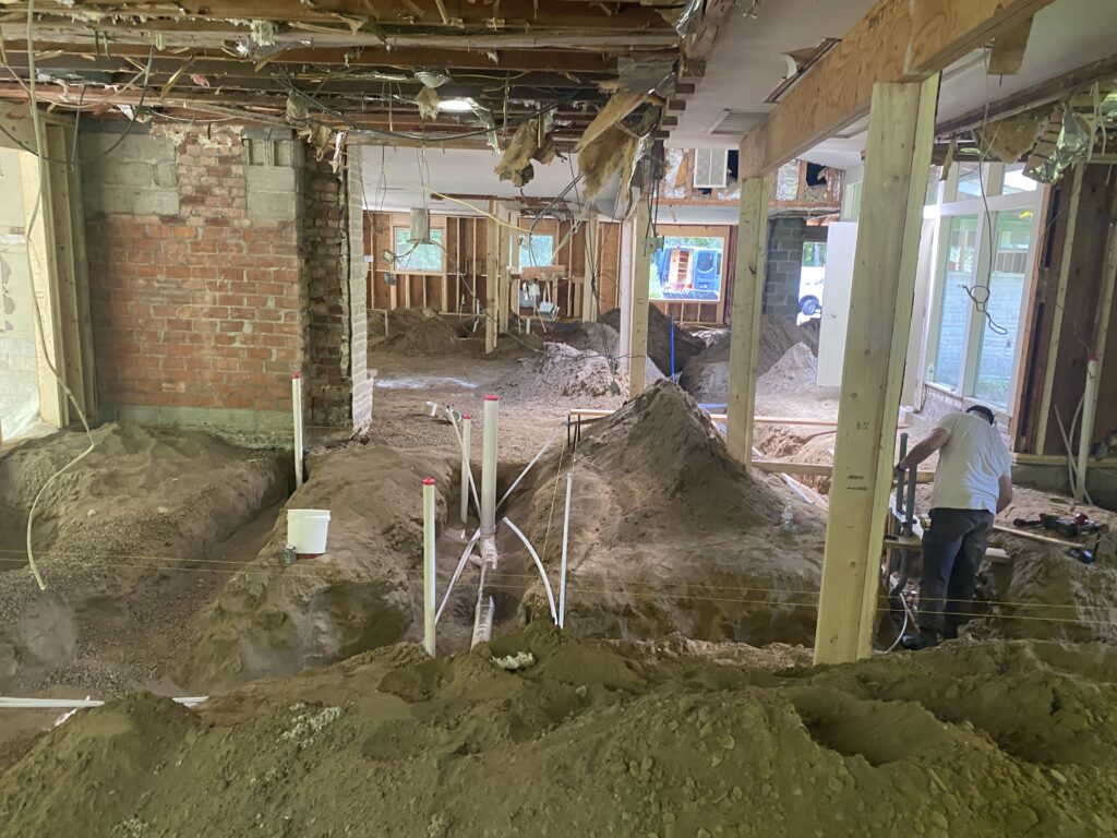Interior demolition scene with exposed ceiling joists, a partially exposed brick chimney, and mounded dirt for new plumbing trenches