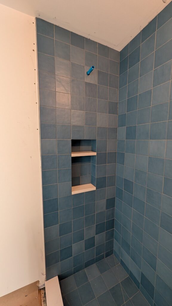 Newly tiled shower stall in progress with blue square wall tile, a recessed two-shelf niche, and a capped shower valve